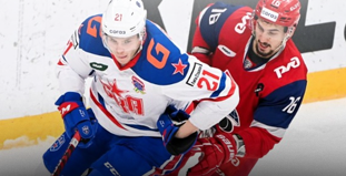 На изображении два хоккеиста в форме разных команд борются за шайбу на льду. Игрок в белой форме с синими и красными элементами соперничает с игроком в красной форме. На заднем плане видна часть рекламного баннера